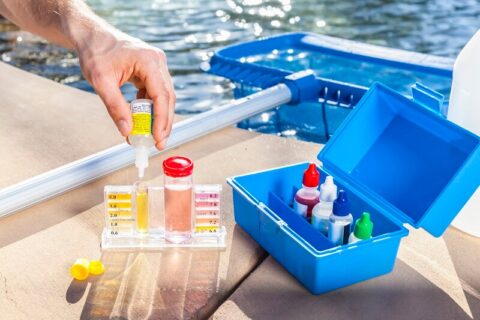 Poolside water testing setup with a chemical test kit showing yellow and pink liquids, a hand holding a yellow testing bottle above it, and a pool in the background.