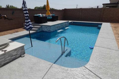 Rectangular swimming pool in a small backyard with a gray poolside surface, raised seating area featuring colorful cushions, and a privacy-ensuring brick wall.