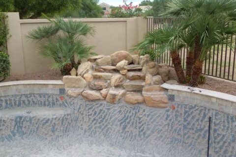Empty swimming pool under renovation with textured gray and blue tiles, decorative rock formation, and palm trees in a backyard setting.