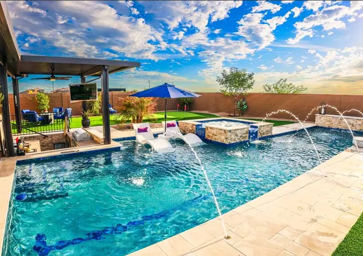 Luxurious backyard pool area in Phoenix, AZ, with a rectangular swimming pool, water jets, built-in lounge chairs, a hot tub with a waterfall feature, and a shaded seating area.